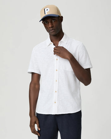 Man wearing a white short-sleeve paige shirt and beige cap with a logo on a plain background