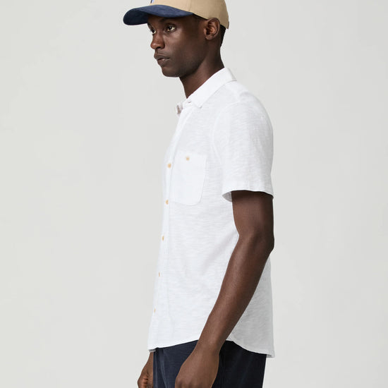Man wearing a white short-sleeve paige shirt and beige cap with a logo on a plain background side view