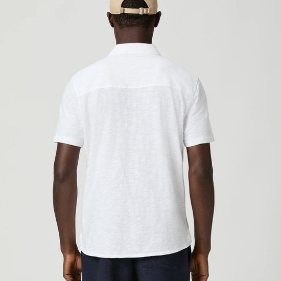 Man wearing a white short-sleeve paige shirt and beige cap with a logo on a plain background rear view