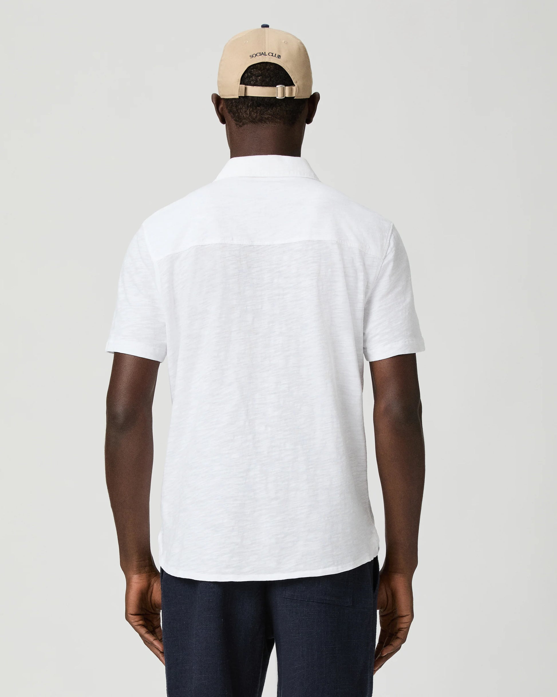 Man wearing a white short-sleeve paige shirt and beige cap with a logo on a plain background rear view