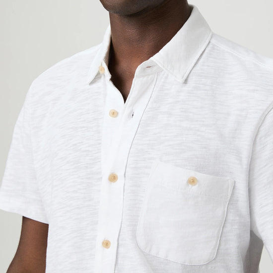 Man wearing a white short-sleeve paige shirt and beige cap with a logo on a plain background close up