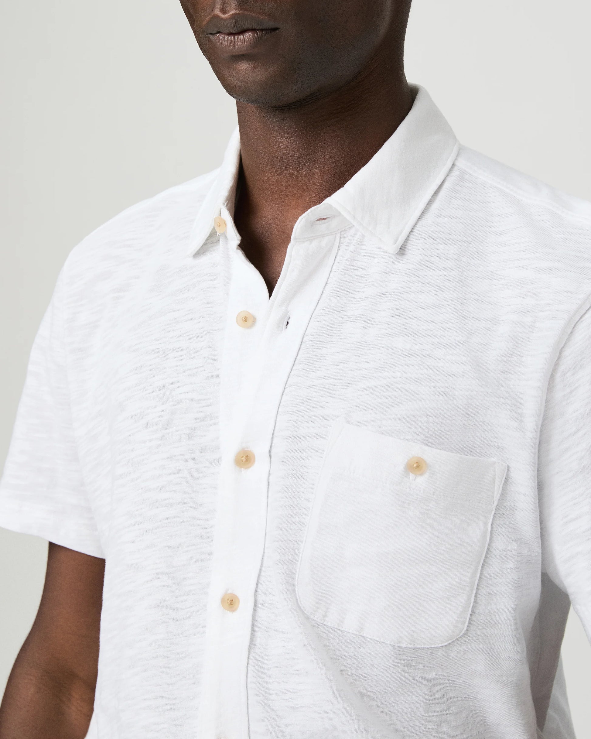 Man wearing a white short-sleeve paige shirt and beige cap with a logo on a plain background close up