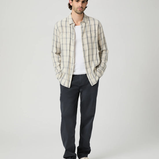 A man stands against a light gray studio background wearing a cream and gray plaid button-up shirt over a white t-shirt, dark charcoal pants, and off-white sneakers.