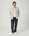 A man stands against a light gray studio background wearing a cream and gray plaid button-up shirt over a white t-shirt, dark charcoal pants, and off-white sneakers.