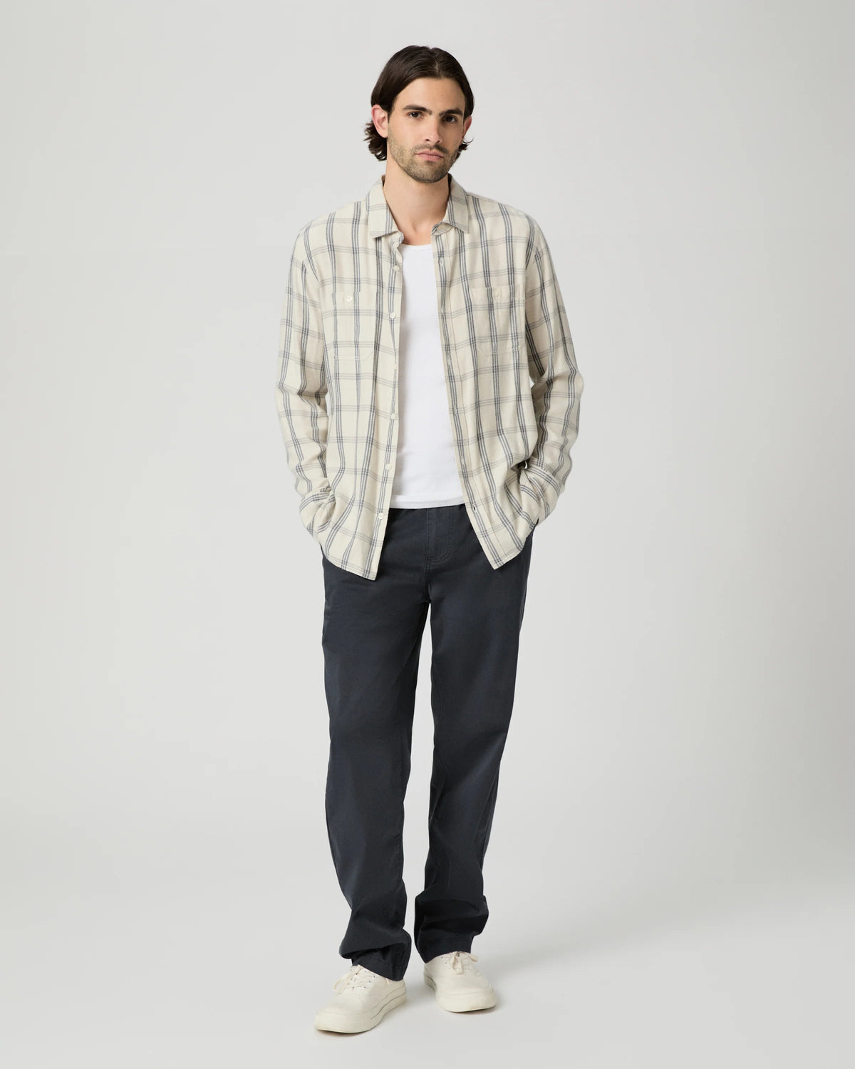 A man stands against a light gray studio background wearing a cream and gray plaid button-up shirt over a white t-shirt, dark charcoal pants, and off-white sneakers.