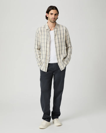A man stands against a light gray studio background wearing a cream and gray plaid button-up shirt over a white t-shirt, dark charcoal pants, and off-white sneakers.