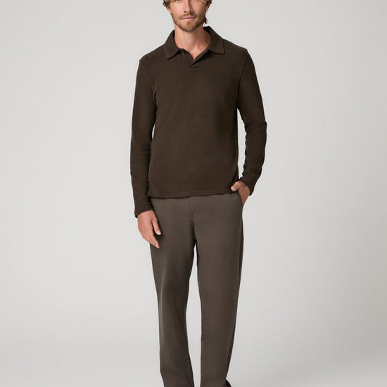 A man modeling a dark brown long-sleeve polo shirt and matching brown trousers against a light gray studio background.