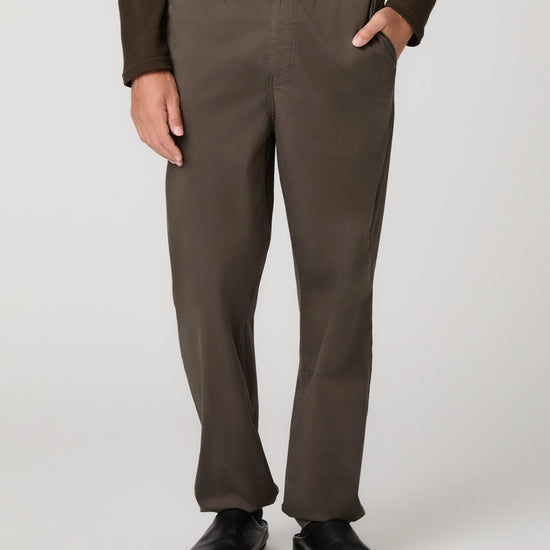 A person wearing dark brown straight-leg trousers, a dark brown long-sleeve top, and black shoes, shown from the waist down against a light gray studio background.