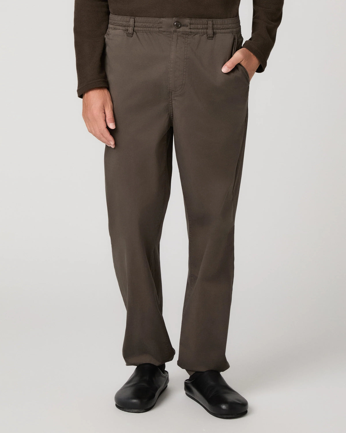 A person wearing dark brown straight-leg trousers, a dark brown long-sleeve top, and black shoes, shown from the waist down against a light gray studio background.