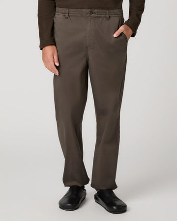 A person wearing dark brown straight-leg trousers, a dark brown long-sleeve top, and black shoes, shown from the waist down against a light gray studio background.