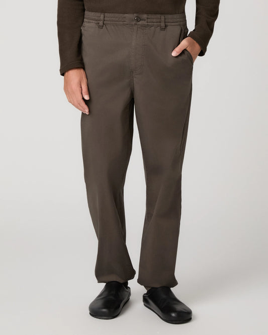 A person wearing dark brown straight-leg trousers, a dark brown long-sleeve top, and black shoes, shown from the waist down against a light gray studio background.