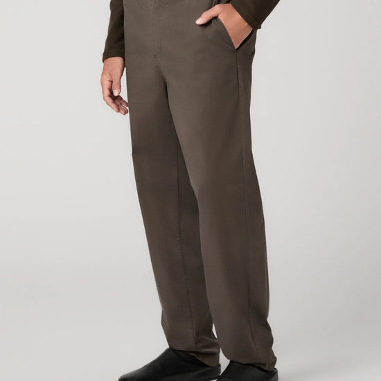 A person wearing dark brown straight-leg trousers, a matching dark brown long-sleeve top, and black shoes, standing against a light gray studio background.