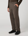 A person wearing dark brown straight-leg trousers, a matching dark brown long-sleeve top, and black shoes, standing against a light gray studio background.