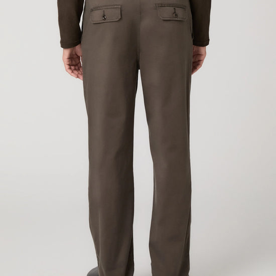 Rear view of a person wearing straight-leg brown trousers with buttoned welt back pockets, paired with a dark brown long-sleeve top and dark shoes, against a light gray studio background.