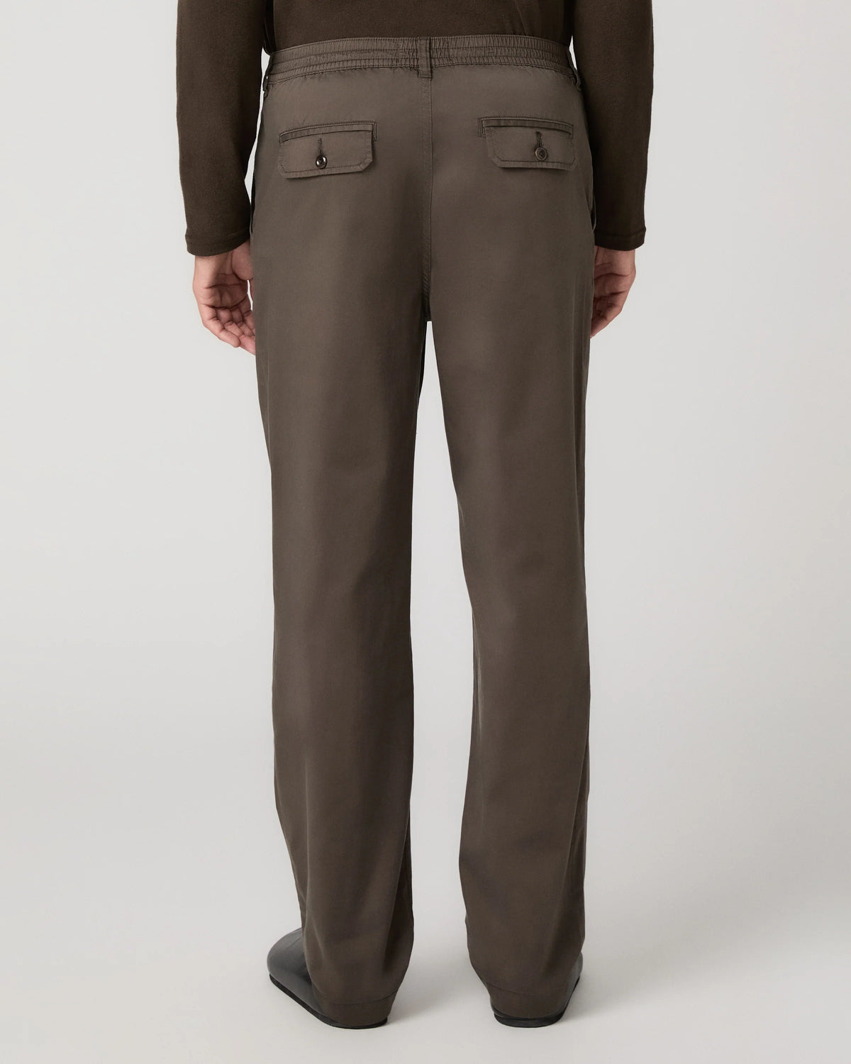 Rear view of a person wearing straight-leg brown trousers with buttoned welt back pockets, paired with a dark brown long-sleeve top and dark shoes, against a light gray studio background.