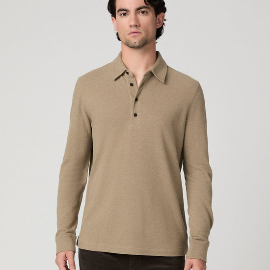 A man wearing a beige long-sleeve polo shirt with a three-button placket, paired with dark pants, standing against a light gray studio background.