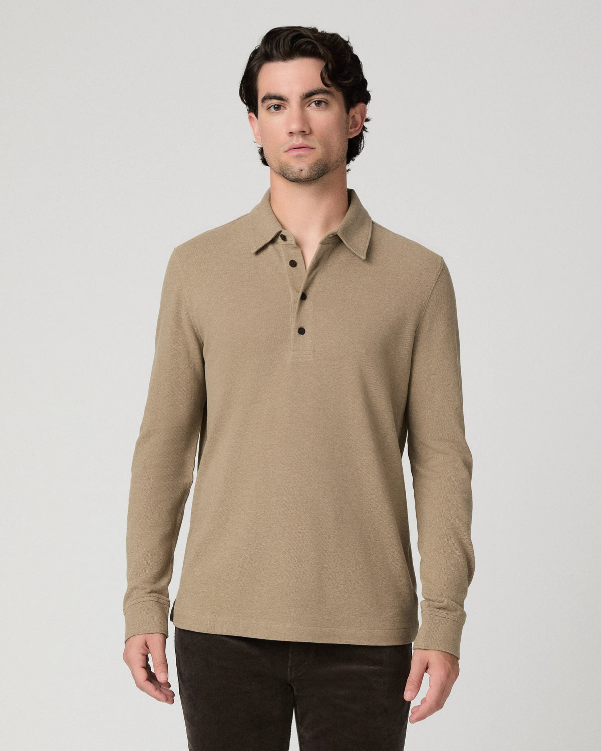 A man wearing a beige long-sleeve polo shirt with a three-button placket, paired with dark pants, standing against a light gray studio background.