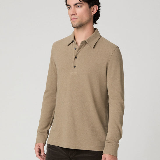 A man wearing a light brown long-sleeve polo shirt with dark brown pants, standing against a light gray studio background.