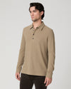 A man wearing a light brown long-sleeve polo shirt with dark brown pants, standing against a light gray studio background.