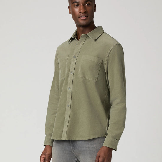 A man wearing a muted sage green long-sleeve button-up shirt and gray jeans, standing against a light gray studio background.