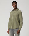 A man wearing a muted sage green long-sleeve button-up shirt and gray jeans, standing against a light gray studio background.