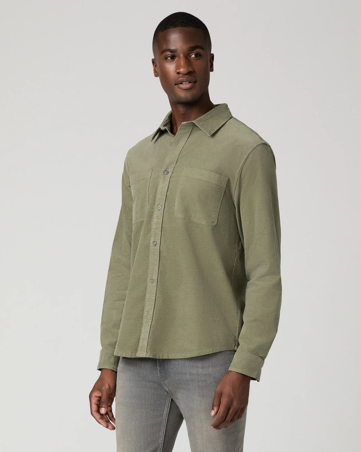 A man wearing a muted sage green long-sleeve button-up shirt and gray jeans, standing against a light gray studio background.