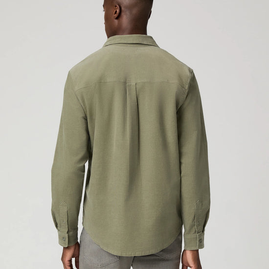 A man viewed from the back wearing a muted sage green long-sleeve button-up shirt and gray trousers, photographed against a plain light gray studio background.
