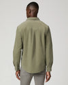 A man viewed from the back wearing a muted sage green long-sleeve button-up shirt and gray trousers, photographed against a plain light gray studio background.