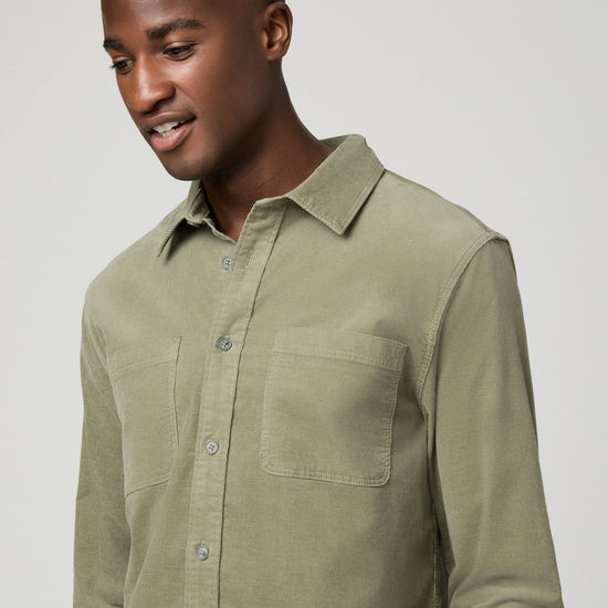 A man wearing a light olive green long-sleeve button-up shirt with two chest pockets, photographed against a neutral light gray background.