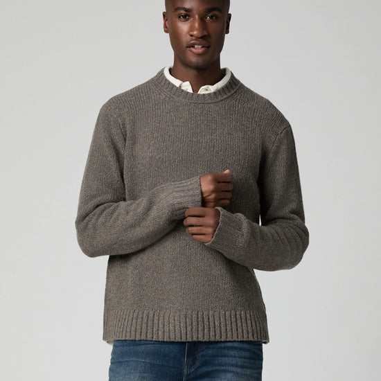 A man wearing a taupe brown ribbed knit sweater over a white collared shirt and blue jeans, standing against a light gray studio background.