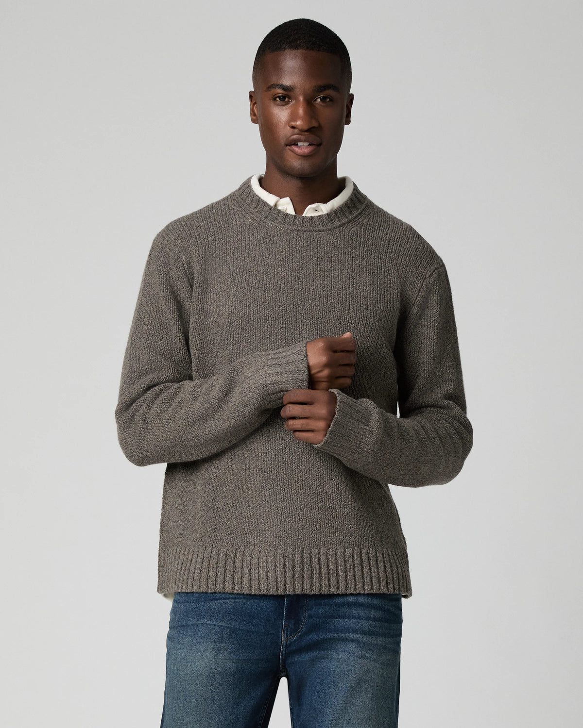 A man wearing a taupe brown ribbed knit sweater over a white collared shirt and blue jeans, standing against a light gray studio background.