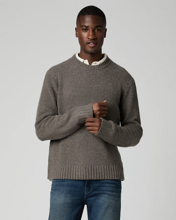 A man wearing a taupe brown ribbed knit sweater over a white collared shirt and blue jeans, standing against a light gray studio background.