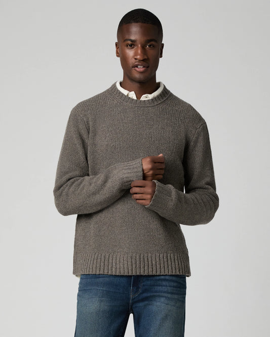 A man wearing a taupe brown ribbed knit sweater over a white collared shirt and blue jeans, standing against a light gray studio background.