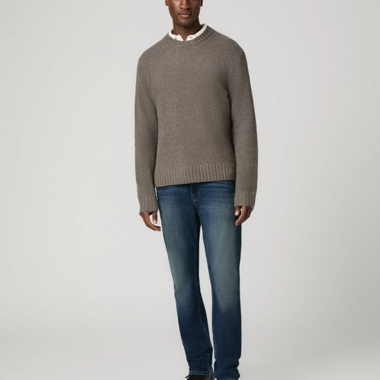 A man modeling a taupe knit crewneck sweater over a white collared shirt, paired with blue jeans and black leather shoes, against a light gray studio background.