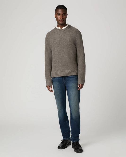 A man modeling a taupe knit crewneck sweater over a white collared shirt, paired with blue jeans and black leather shoes, against a light gray studio background.