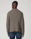 A man viewed from the back wearing a taupe brown ribbed knit sweater and blue jeans, set against a plain light gray studio background.