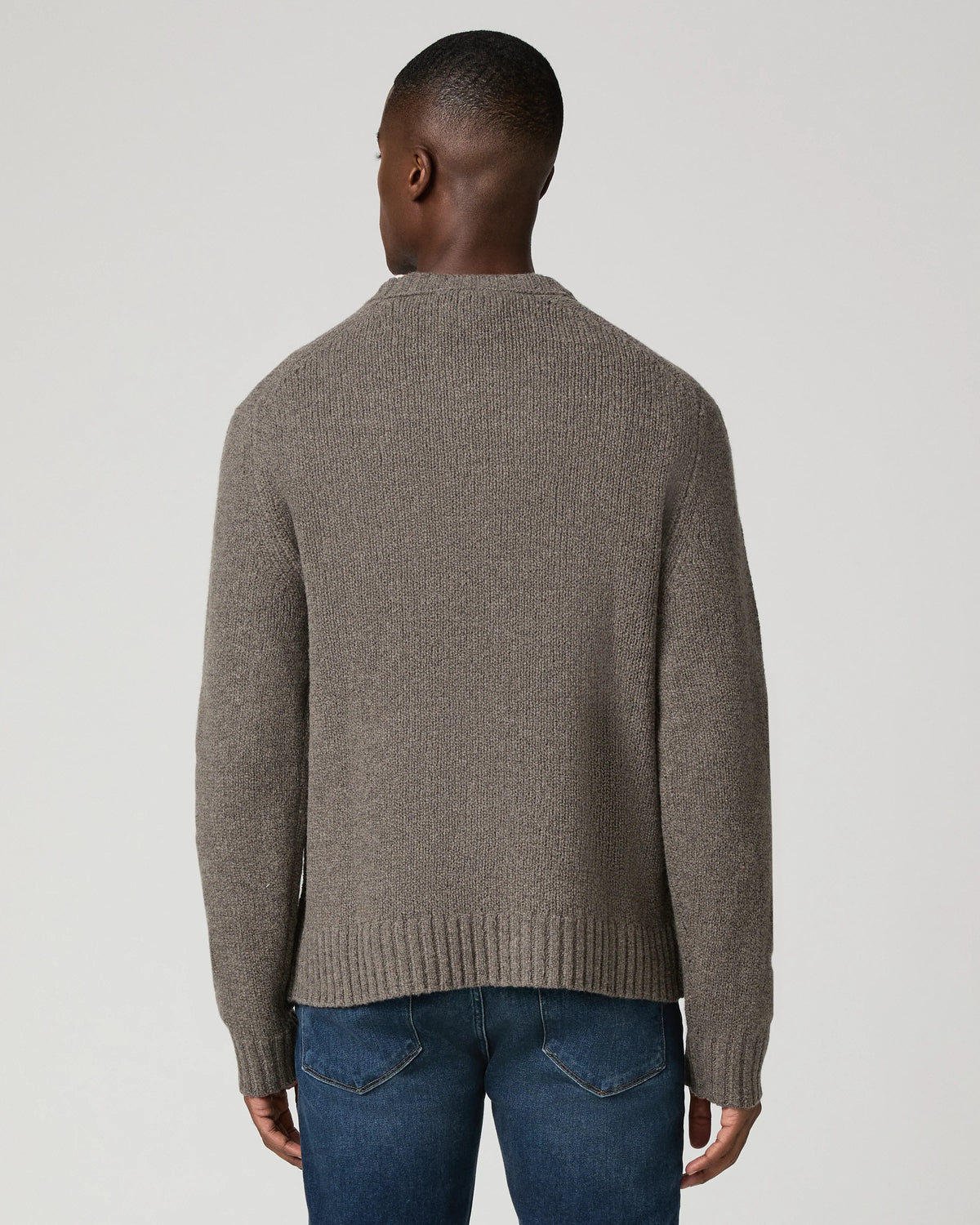A man viewed from the back wearing a taupe brown ribbed knit sweater and blue jeans, set against a plain light gray studio background.