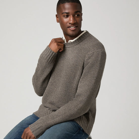 A man wearing a taupe brown ribbed knit sweater over a white collared shirt and blue jeans, sitting on a stool against a light gray background.