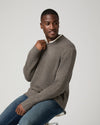 A man wearing a taupe brown ribbed knit sweater over a white collared shirt and blue jeans, sitting on a stool against a light gray background.