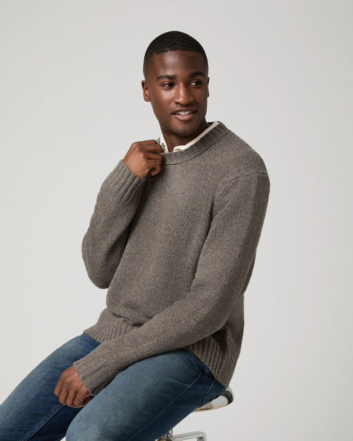 A man wearing a taupe brown ribbed knit sweater over a white collared shirt and blue jeans, sitting on a stool against a light gray background.