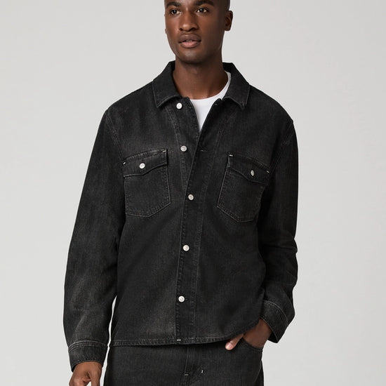 A man wearing a dark washed denim button-up shirt jacket over a white t-shirt and dark jeans, standing against a light gray studio background.