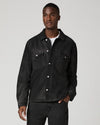 A man wearing a dark washed denim button-up shirt jacket over a white t-shirt and dark jeans, standing against a light gray studio background.