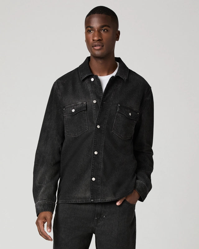 A man wearing a dark washed denim button-up shirt jacket over a white t-shirt and dark jeans, standing against a light gray studio background.