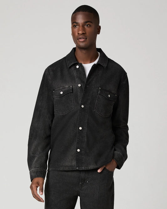 A man wearing a dark washed denim button-up shirt jacket over a white t-shirt and dark jeans, standing against a light gray studio background.
