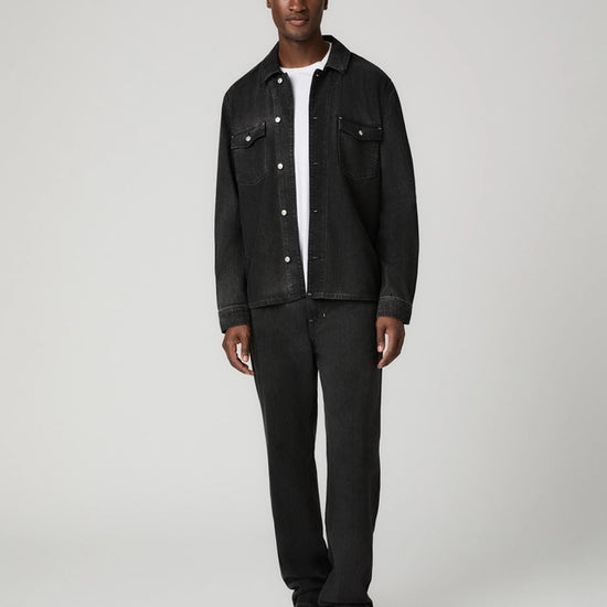 A man modeling a black denim jacket and matching black jeans over a white t-shirt, standing against a light gray studio background.