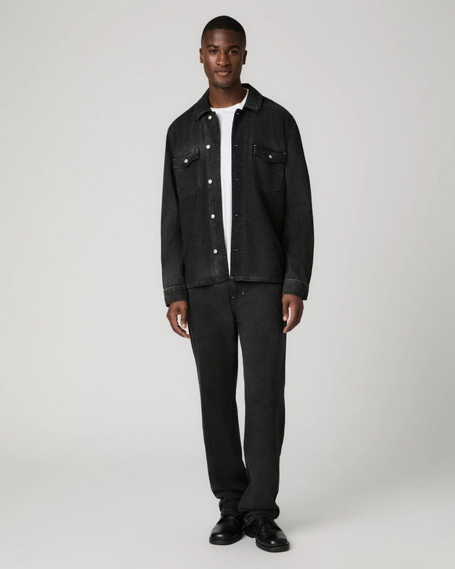 A man modeling a black denim jacket and matching black jeans over a white t-shirt, standing against a light gray studio background.