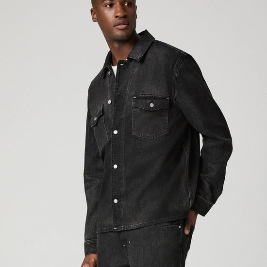 A man wearing a dark washed denim button-up shirt jacket and matching dark jeans, standing against a light gray studio background.
