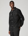 A man wearing a dark washed denim button-up shirt jacket and matching dark jeans, standing against a light gray studio background.