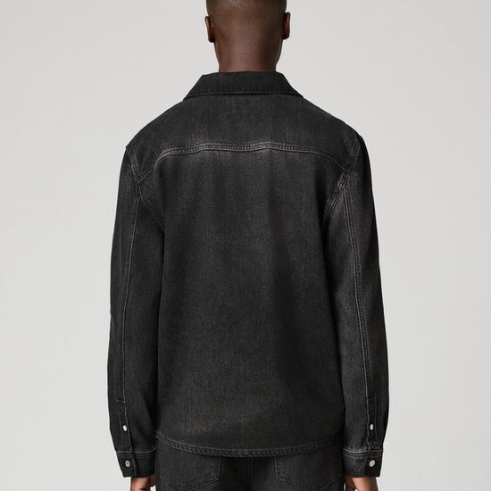 A man viewed from the back wearing a dark washed denim jacket and matching dark jeans against a light gray studio background.