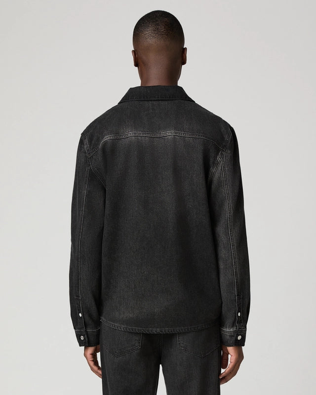 A man viewed from the back wearing a dark washed denim jacket and matching dark jeans against a light gray studio background.